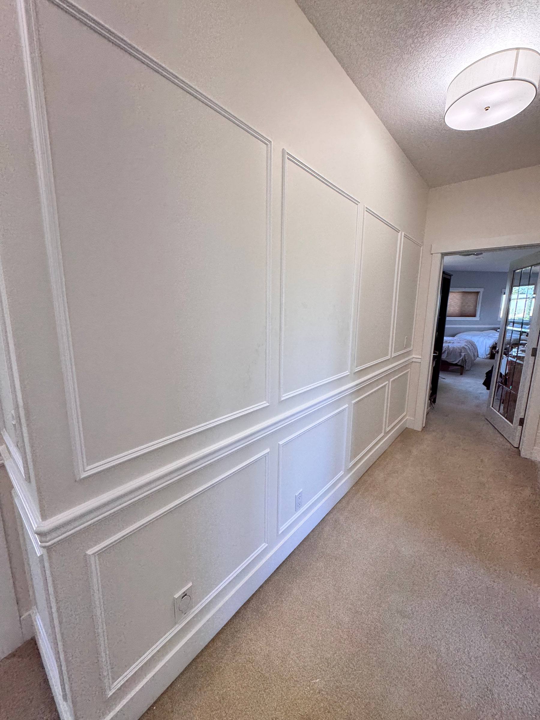 Hallway lined with tall picture-frame panels and wainscoting leading to a bedroom